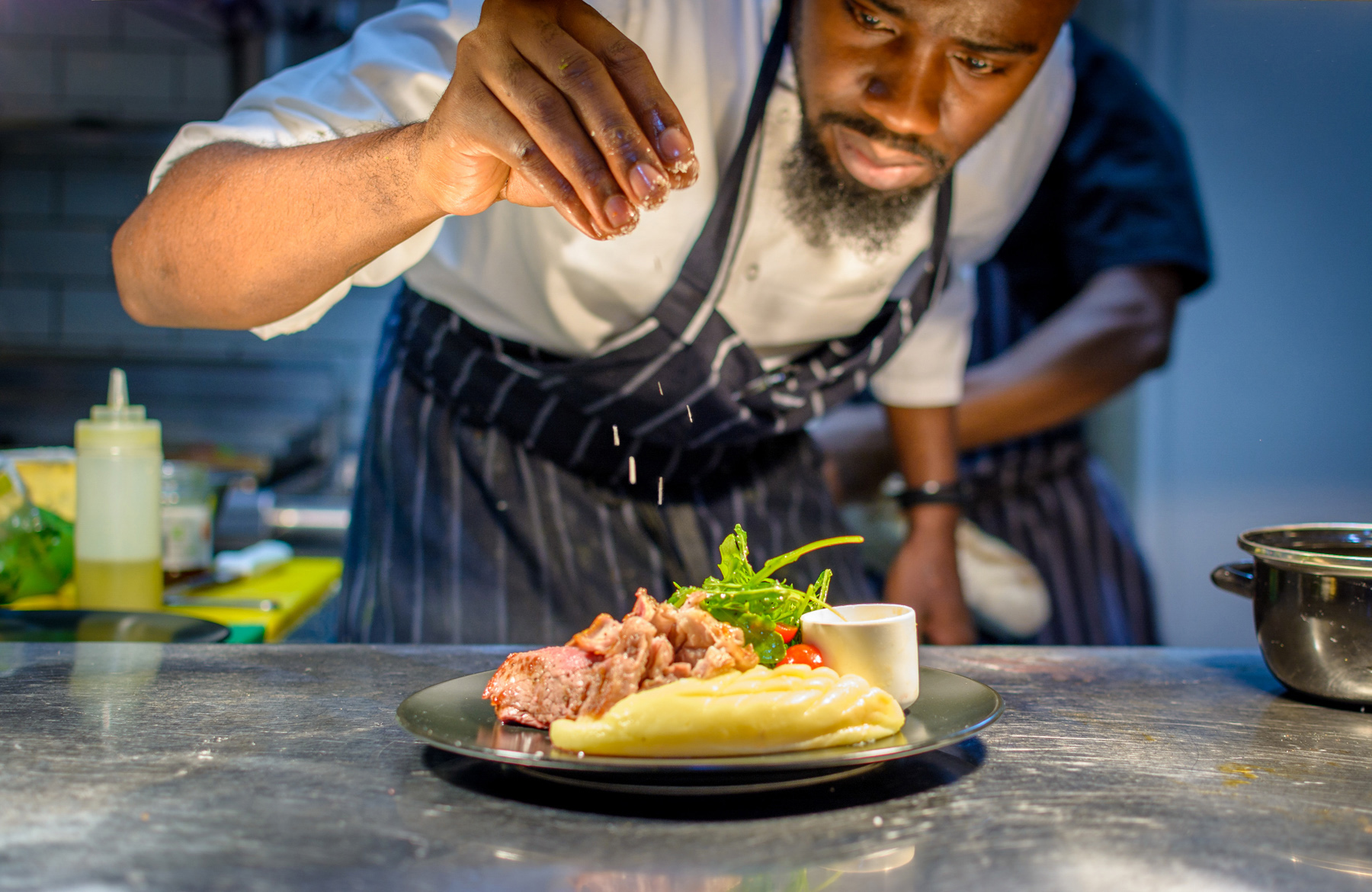 An image showing a chef seasoning a plate of food in a commercial kitchen.