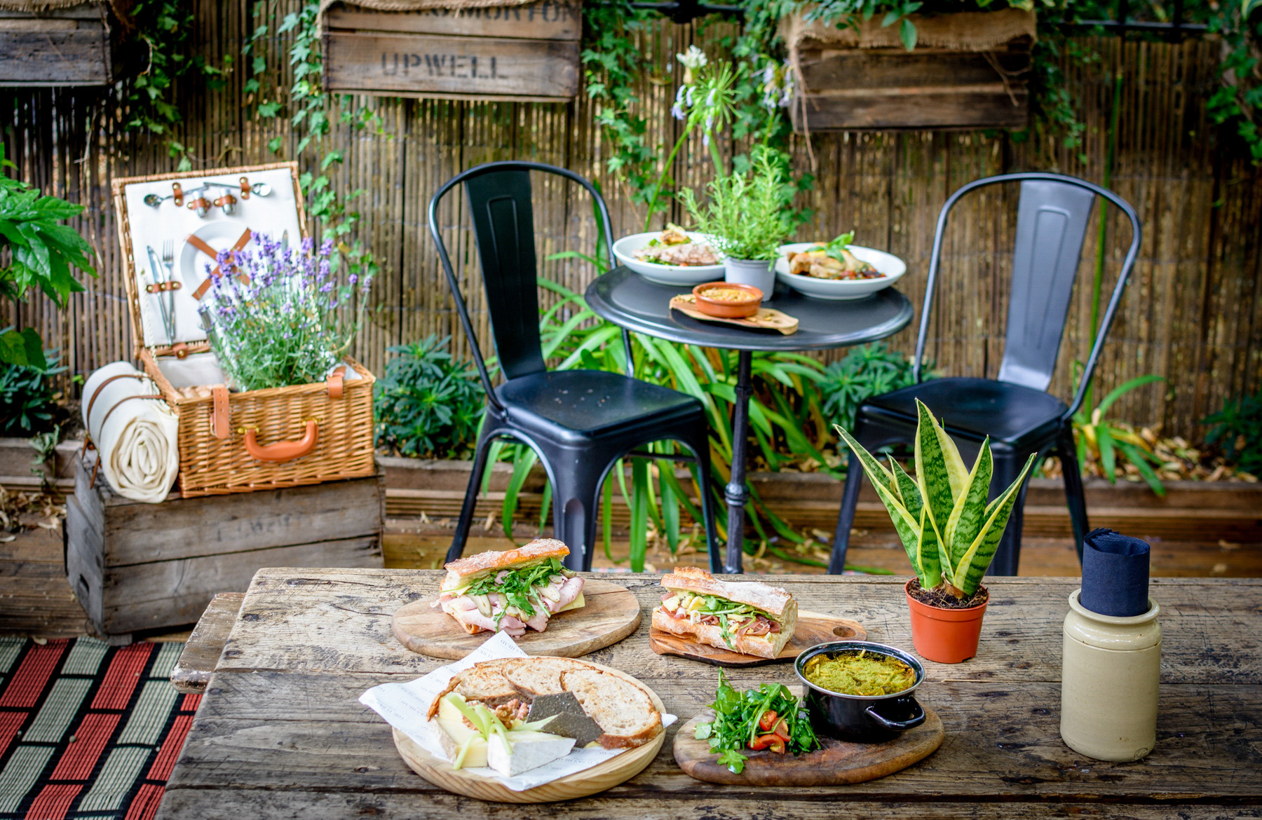 An image showing various food plates on an outside table with plants and chairs.